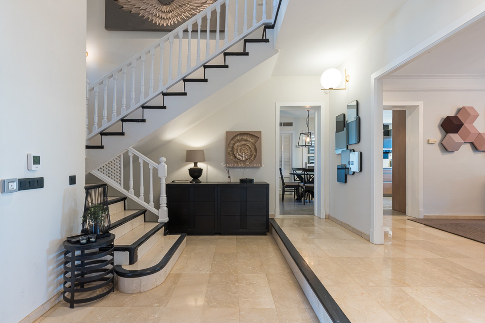 Hall de entrada de lujo con suelos de mármol crema, escalera blanca con detalles negros y decoración contemporánea.
