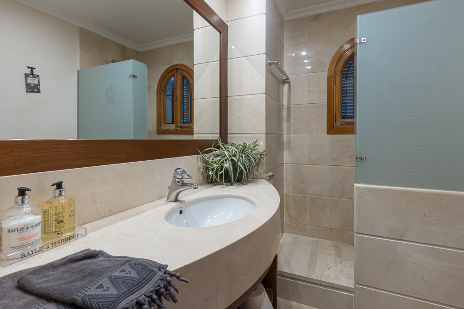 Baño con lavabo de mármol crema, gran espejo con marco de madera y ducha con mampara de cristal ácido.