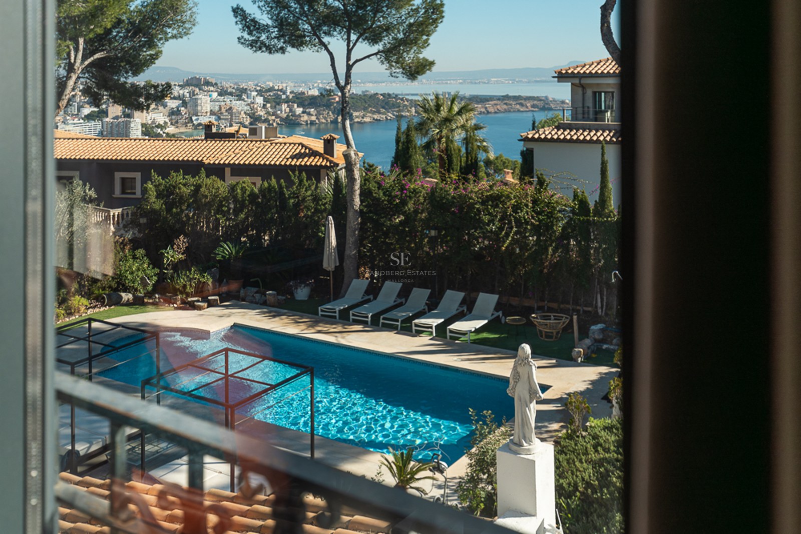 Vista desde una planta superior de una piscina turquesa, tumbonas y la bahía mediterránea al fondo bajo un cielo despejado.