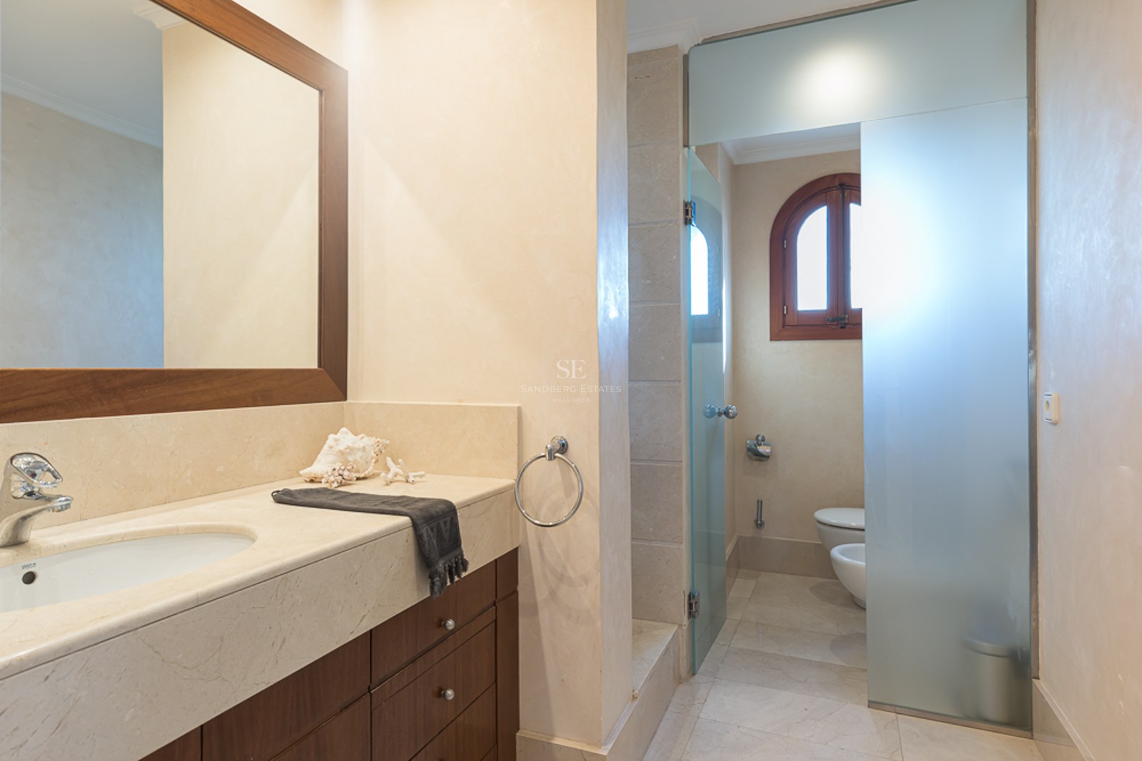 Baño moderno con encimera de mármol crema, muebles de madera oscura y panel de cristal translúcido.