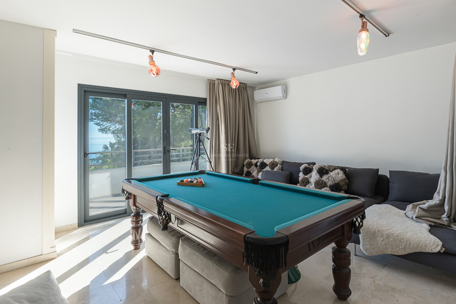 Sala de juegos con mesa de billar de madera, sofás grises y puertas correderas de cristal con vistas al exterior.