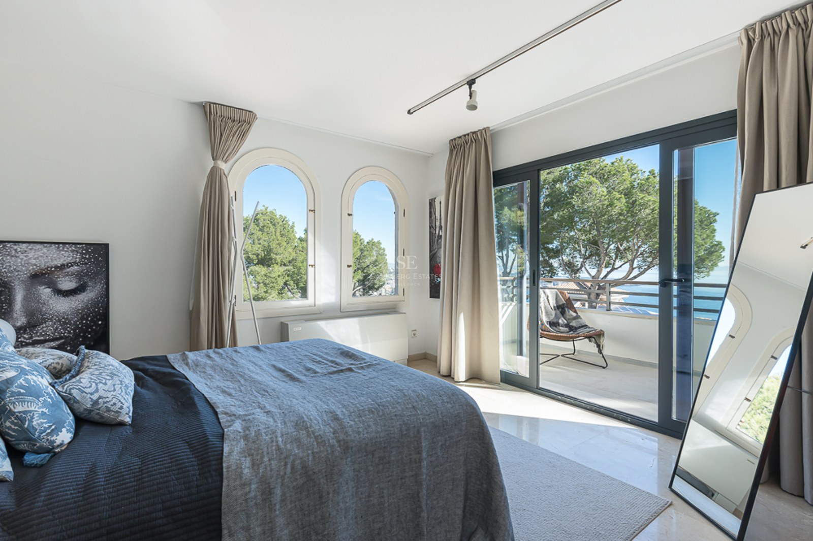 Dormitorio luminoso con ventanas en arco y acceso a terraza con vistas al Mediterráneo y pinos.