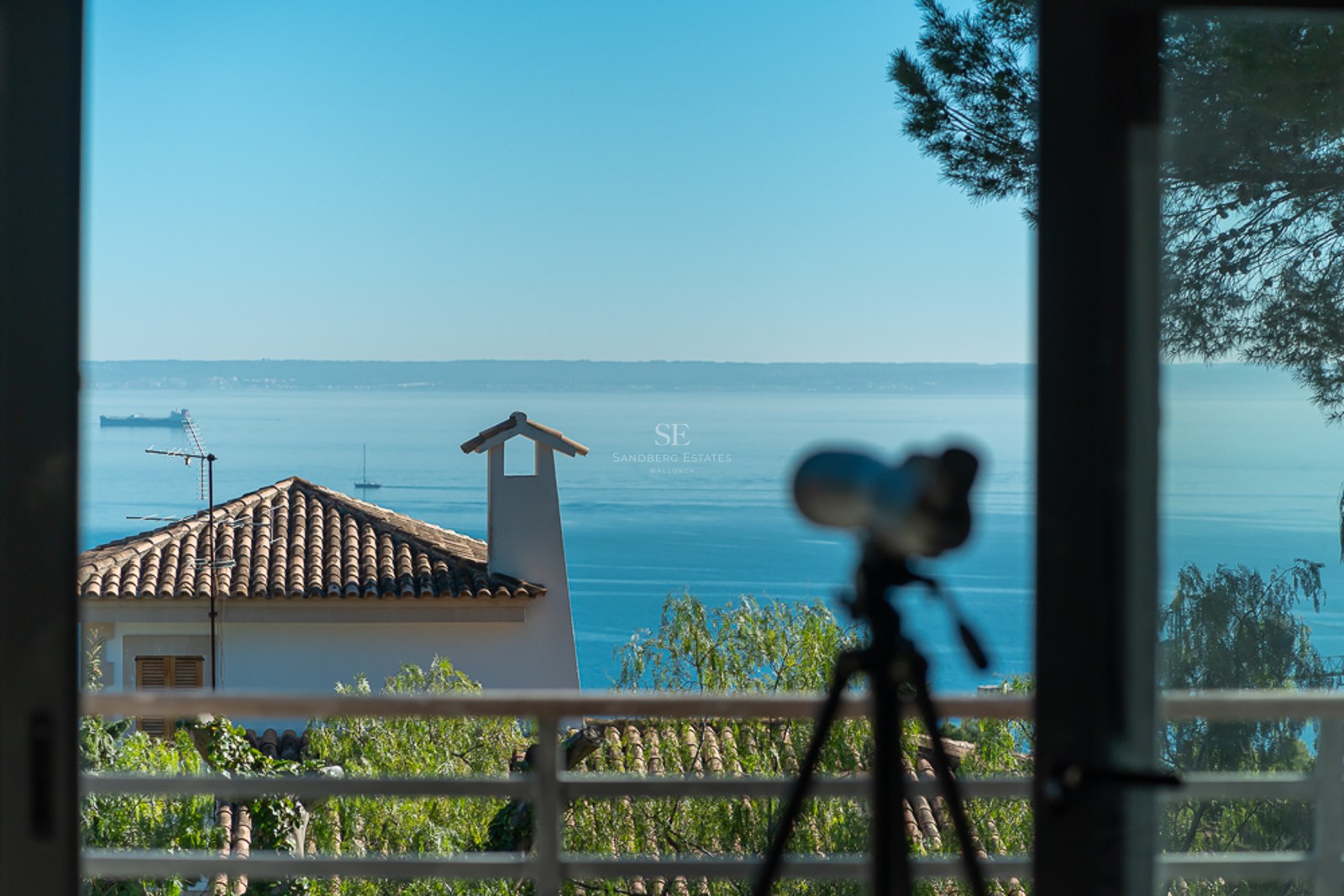 Vista desde el interior hacia tejados tradicionales y vegetación con el mar azul y el horizonte al fondo.