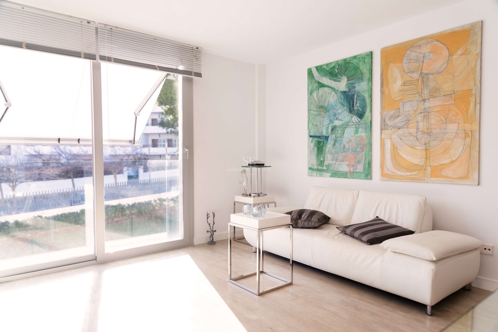 Salon lumineux avec canapé en cuir blanc, deux grands tableaux abstraits et baies vitrées toute hauteur.