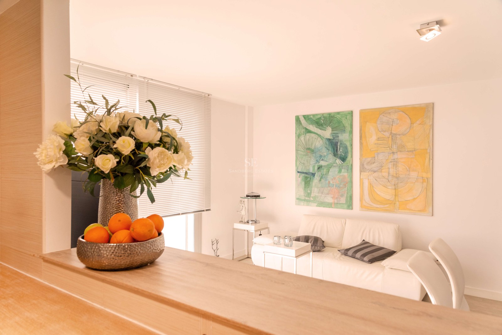 Salon lumineux avec canapé en cuir blanc, peintures abstraites et comptoir en bois avec fleurs.