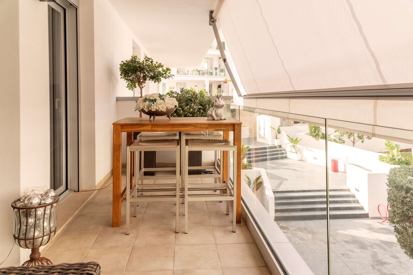 Terrasse moderne avec table haute en bois, tabourets, store blanc et garde-corps en verre donnant sur la cour.