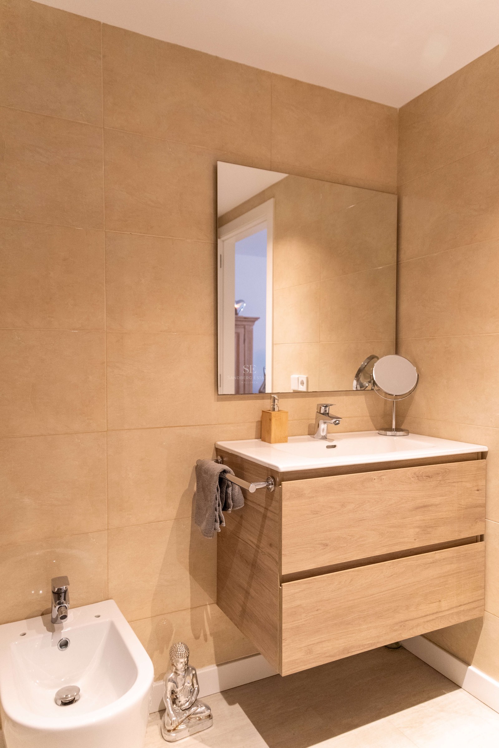 Salle de bain moderne avec meuble vasque suspendu en chêne, lavabo en céramique, grand miroir et carrelage beige.