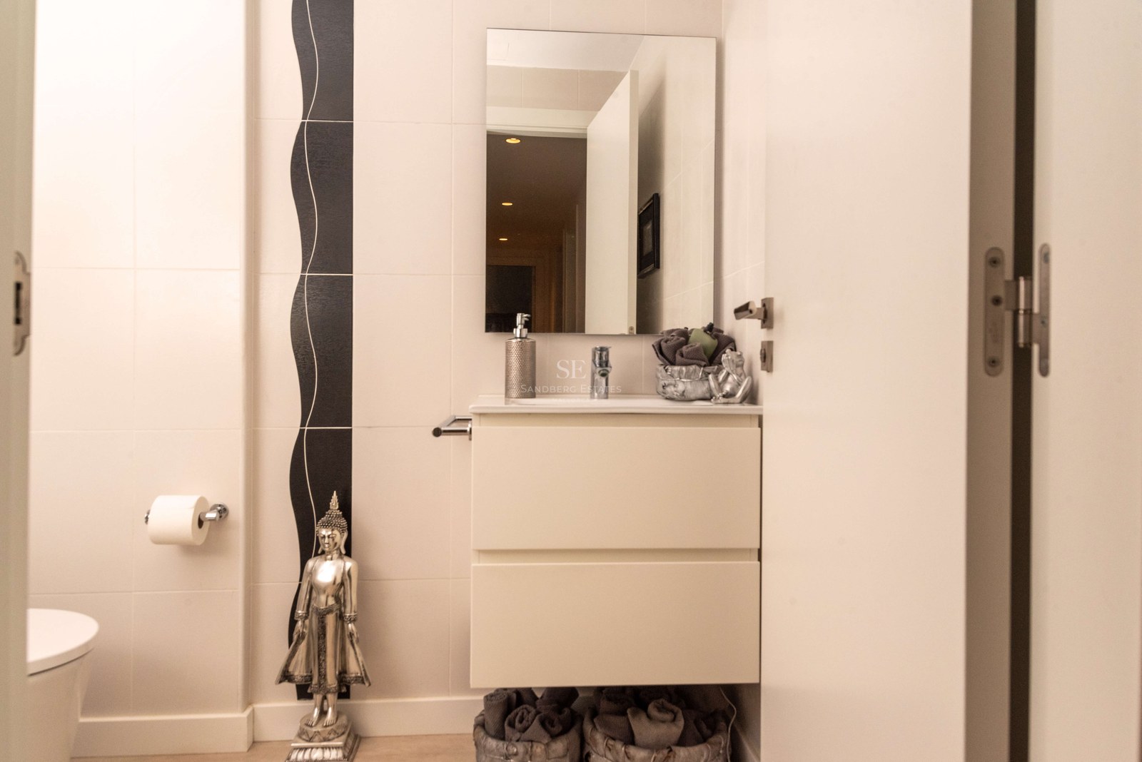 Salle de bains crème avec vanité suspendue blanche, miroir rectangulaire et statue de Bouddha argentée.