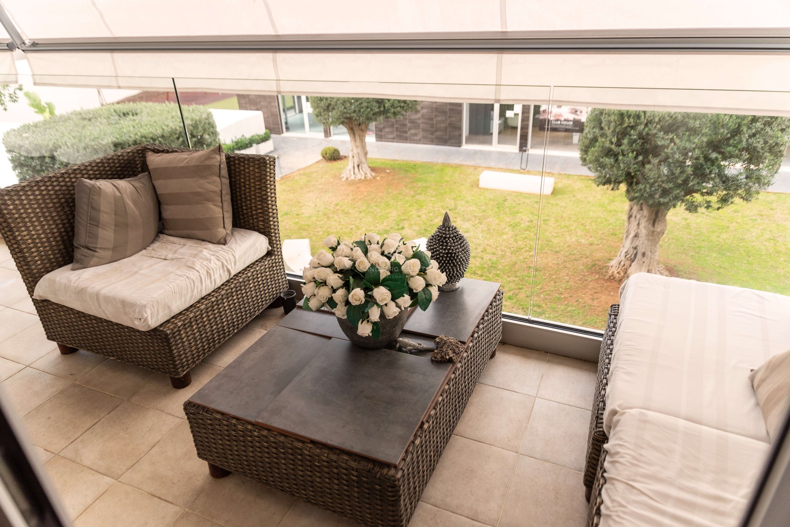 Terrasse couverte avec salon en osier, garde-corps en verre et vue sur un jardin avec oliviers.