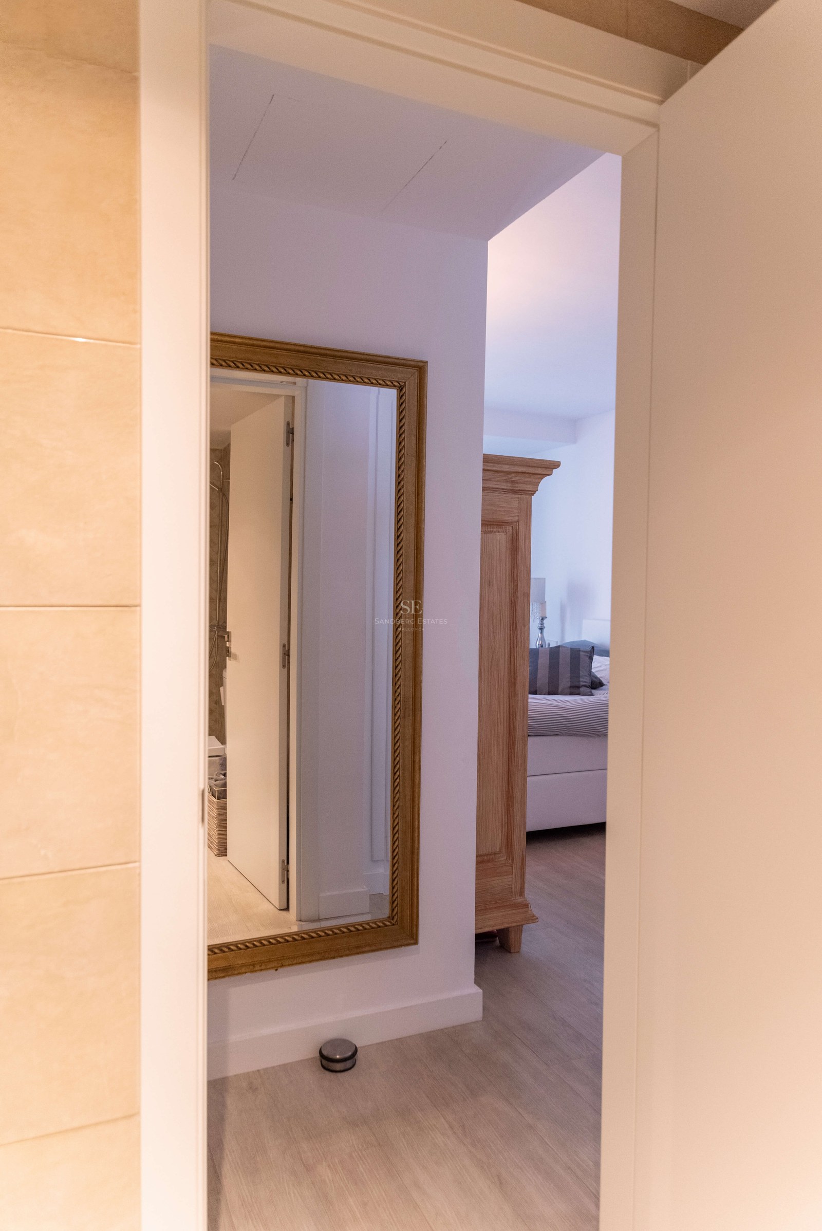 Vue à travers une porte ouverte montrant un couloir avec un miroir doré et une chambre avec armoire en bois.