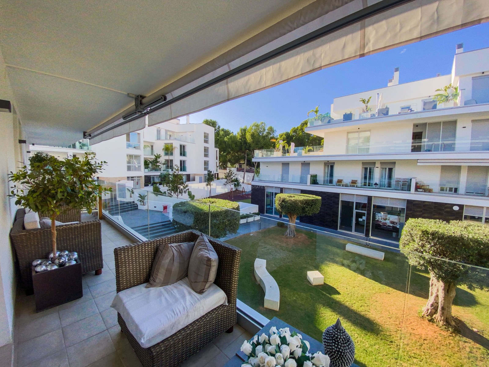 Terrasse avec fauteuils en osier et garde-corps en verre donnant sur un jardin et des immeubles blancs modernes.