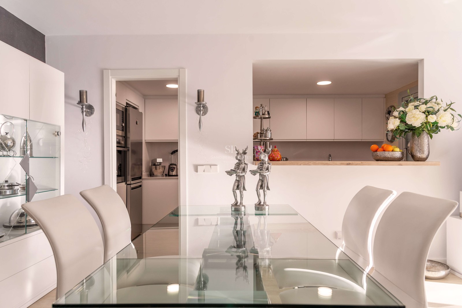 Salle à manger contemporaine avec table en verre, chaises blanches et vue sur une cuisine blanche moderne.