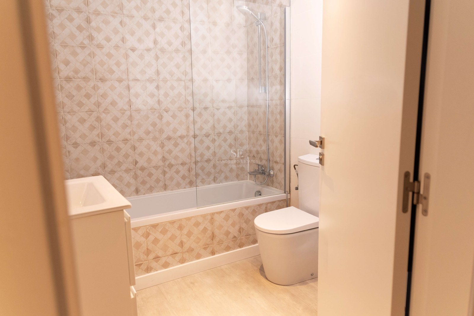 Salle de bain avec carrelage géométrique beige, baignoire avec paroi en verre, toilettes et meuble vasque.