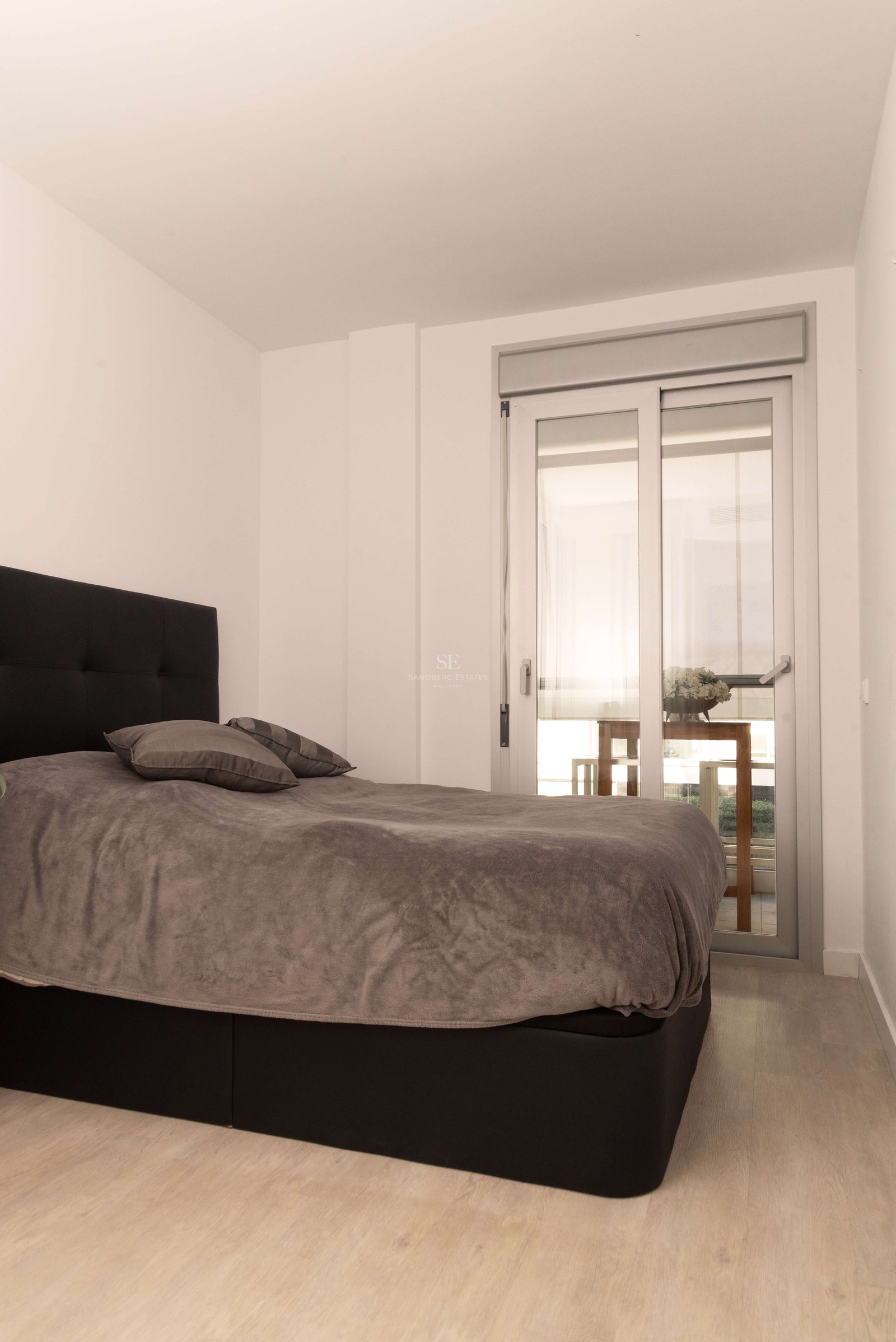 Chambre avec lit gris, tête de lit noire, parquet clair et porte vitrée coulissante menant à une terrasse.
