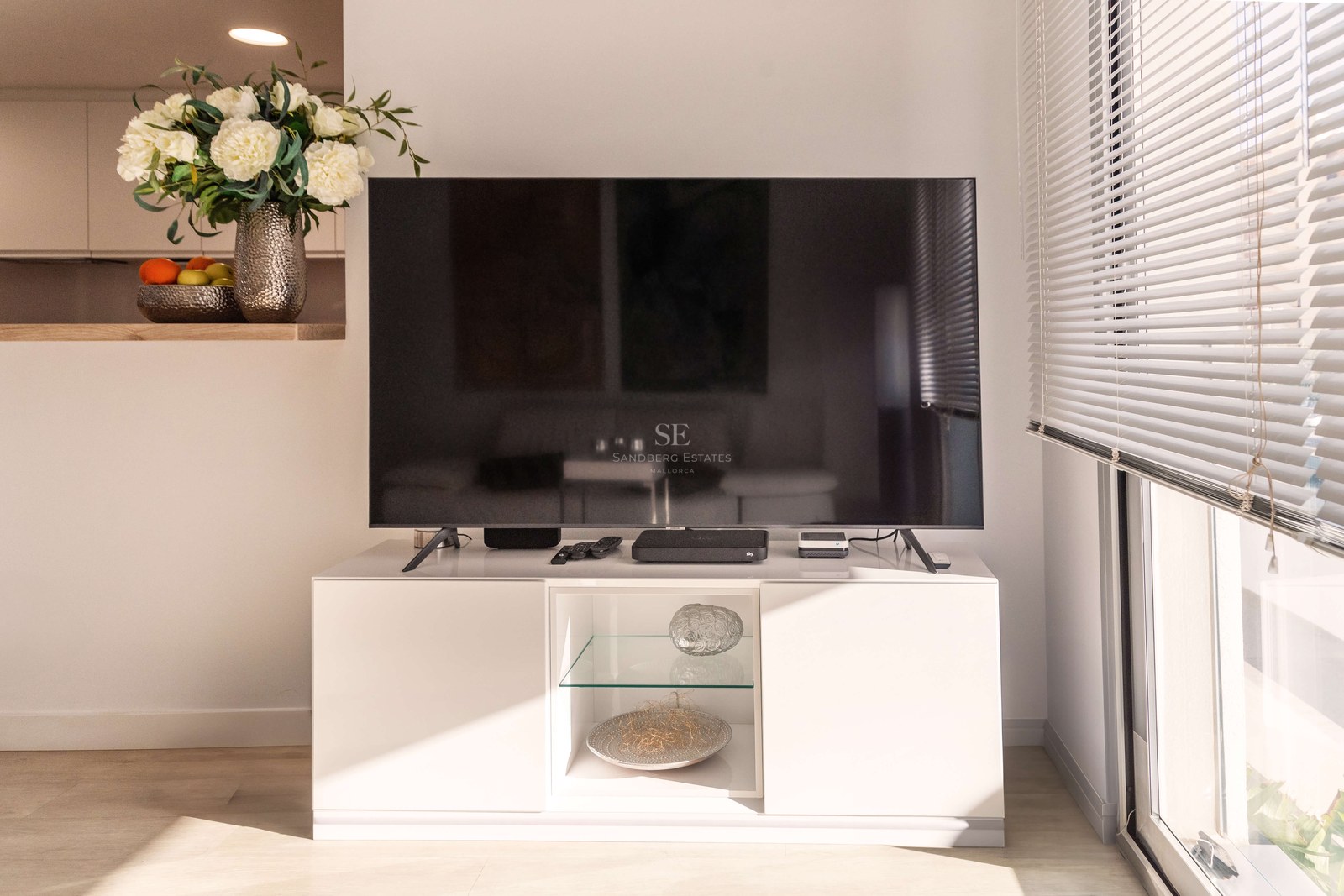 Grand téléviseur sur un buffet blanc minimaliste à côté d'une fenêtre lumineuse avec stores et décor floral.