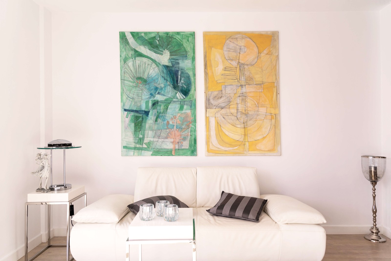 Canapé en cuir blanc avec finitions chromées et deux grandes peintures abstraites verte et jaune sur mur blanc.