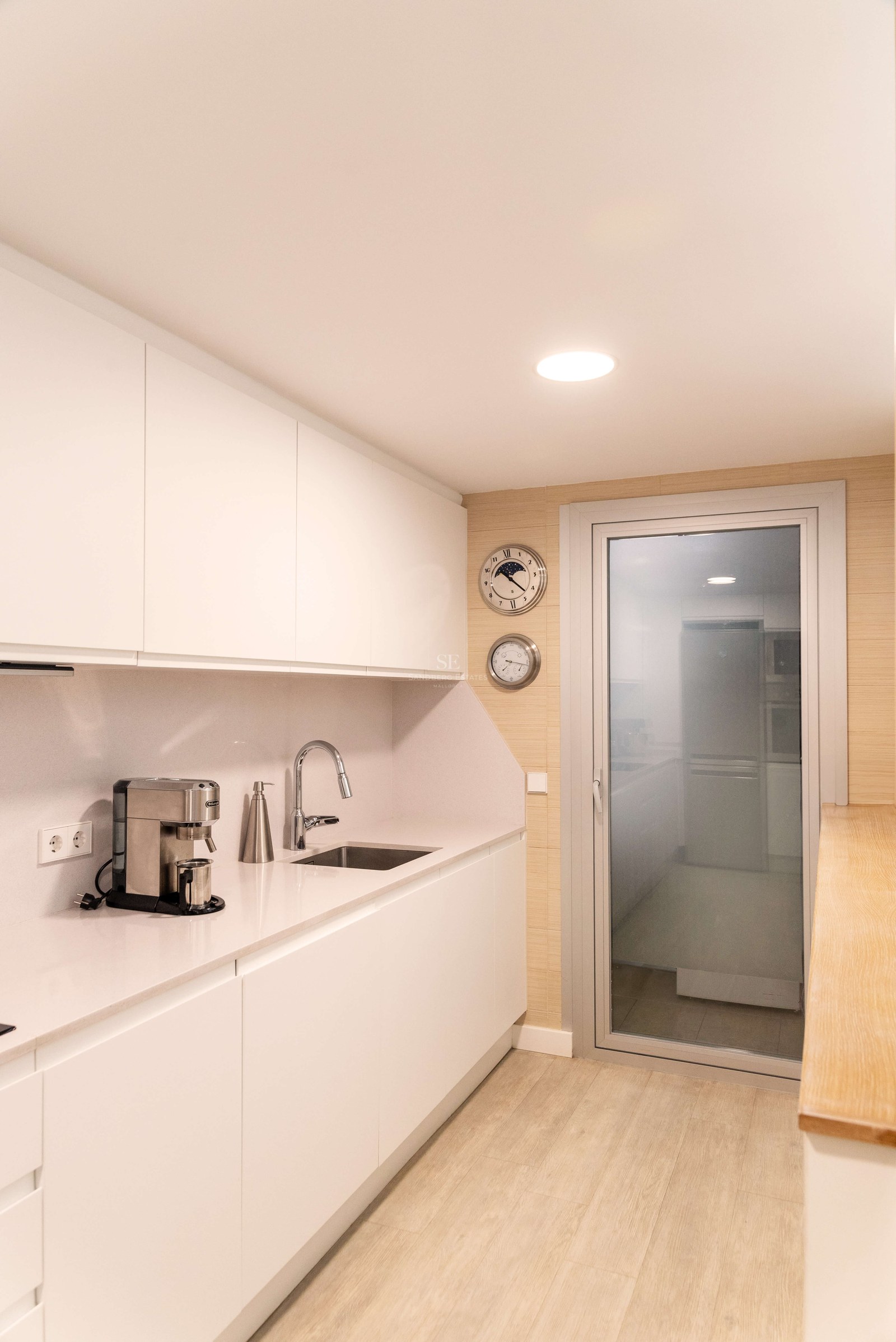 Cuisine en couloir blanche avec placards sans poignées, évier en inox et sol en bois clair.