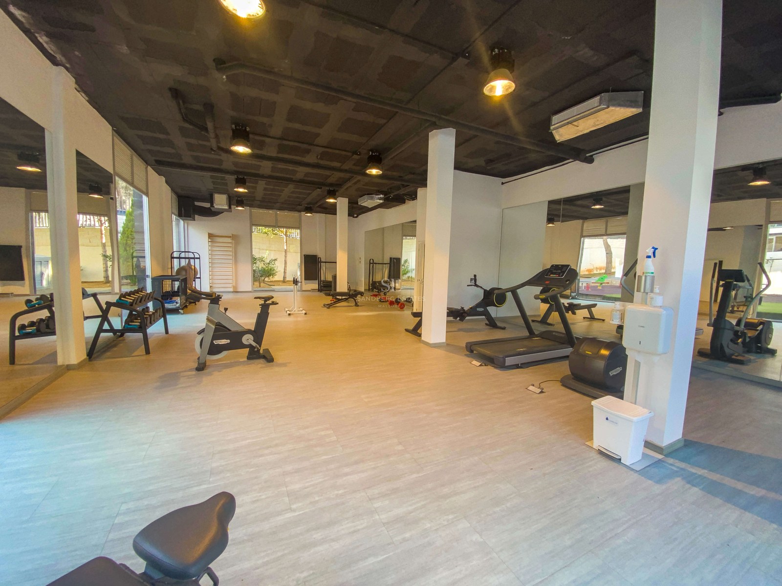 Grande salle de sport intérieure avec équipement Technogym, miroirs toute hauteur et plafond industriel en béton.