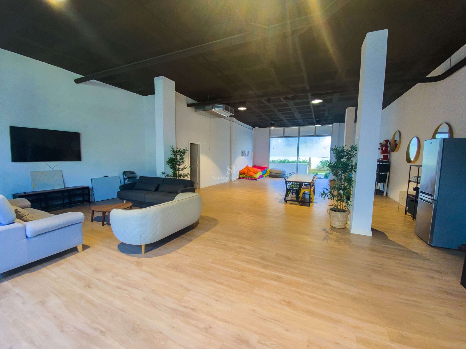 Grande pièce à aire ouverte avec parquet clair, canapés, table à manger, poufs colorés et accès extérieur.
