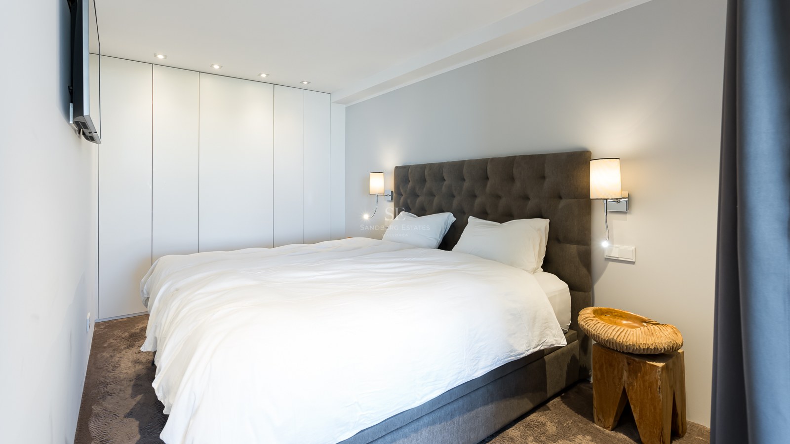 Chambre contemporaine avec lit blanc, tête de lit grise capitonnée, placards blancs et table de chevet en bois brut.