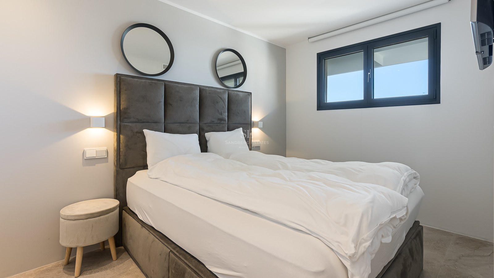 Chambre moderne avec tête de lit en velours gris, miroirs ronds, appliques murales et fenêtre à cadre noir.