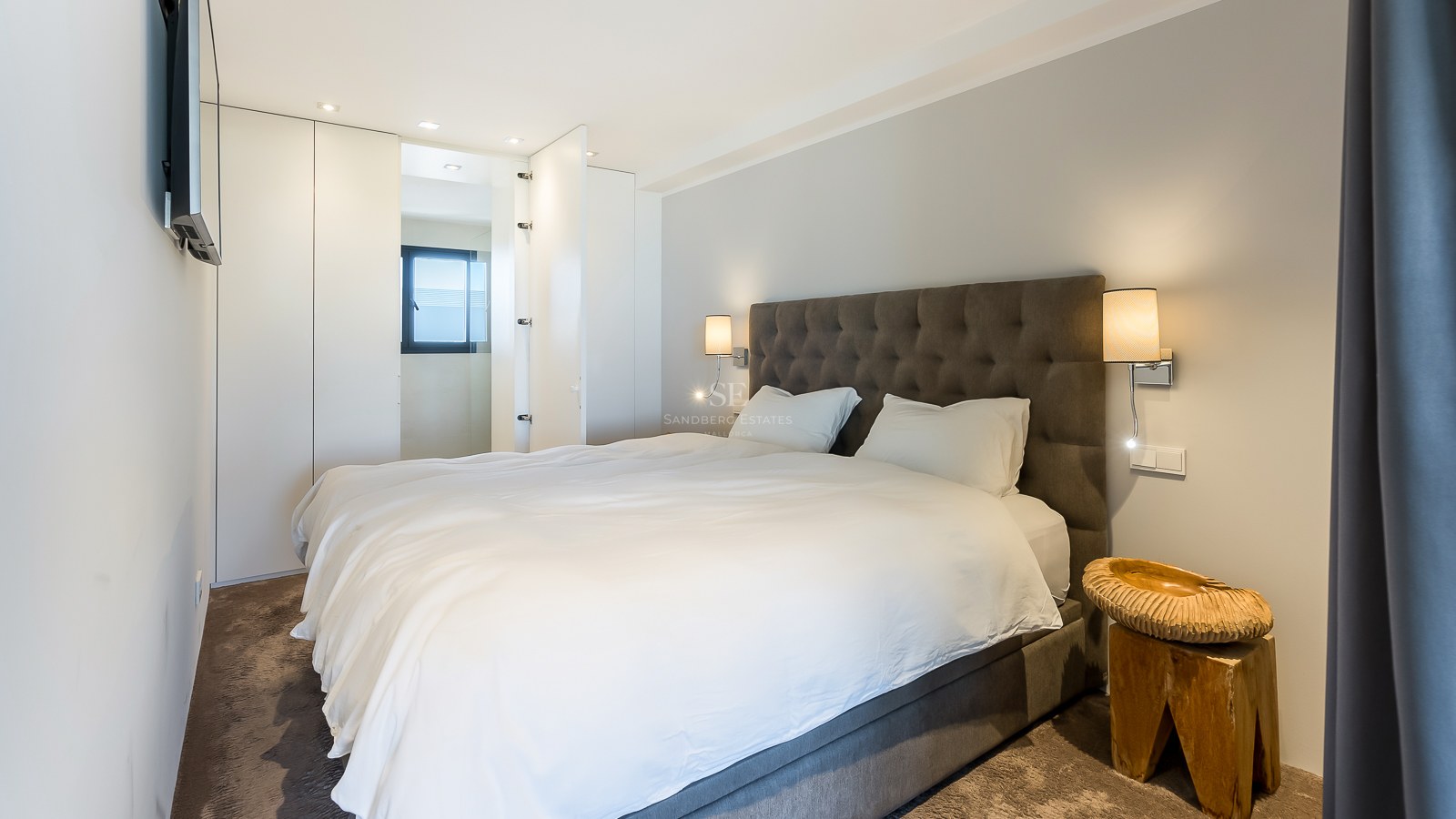 Chambre moderne avec tête de lit capitonnée grise, placards intégrés blancs et table de chevet en bois sculpté.