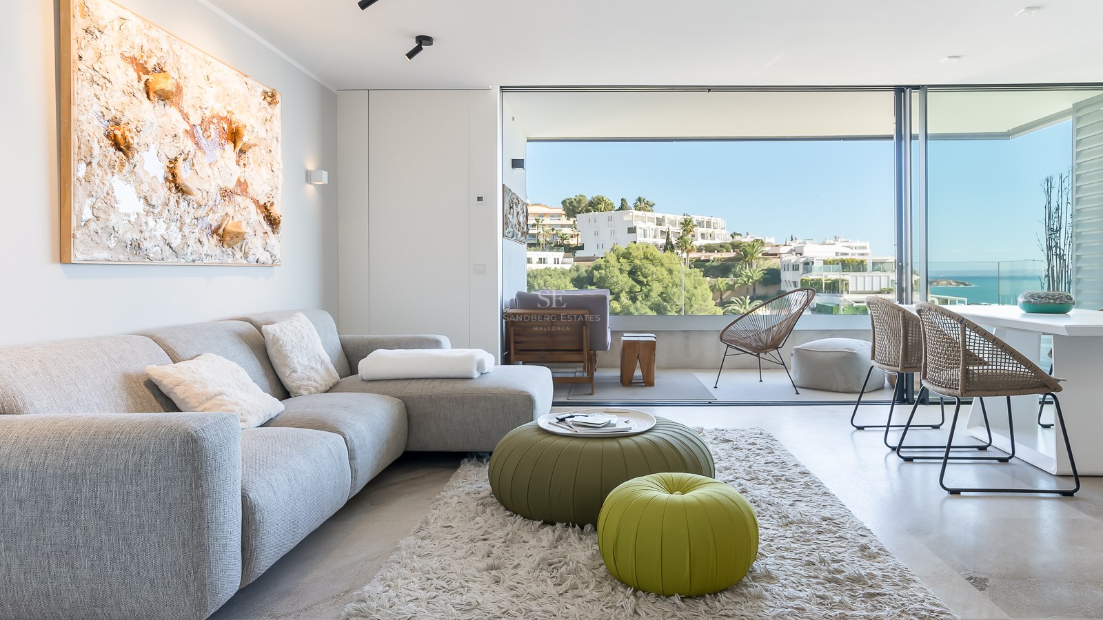 Salon moderne et lumineux avec canapé gris et poufs verts, doté de baies vitrées offrant une vue sur la mer.