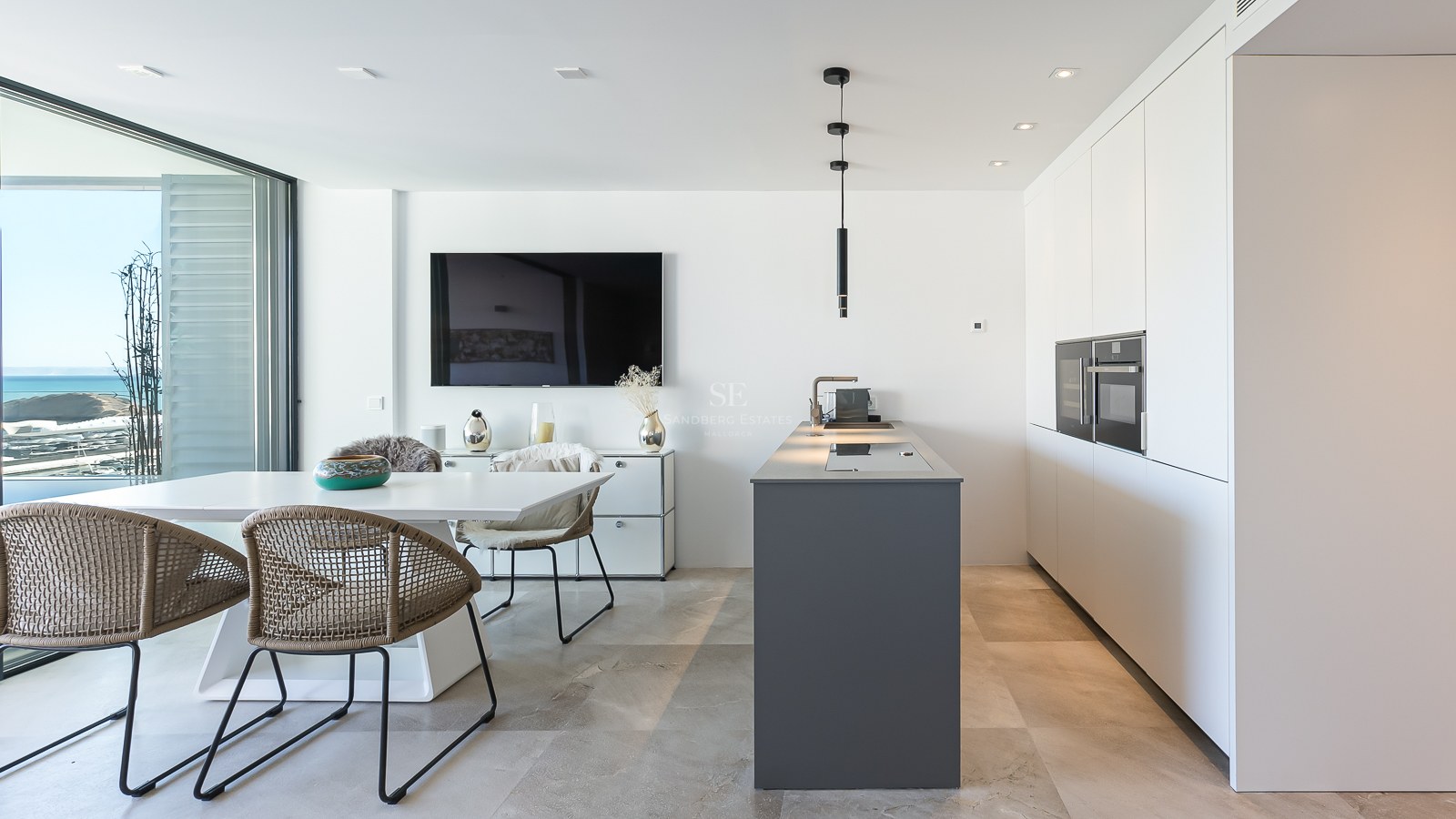 Cuisine blanche moderne avec îlot gris, table à manger et grandes baies vitrées donnant sur la mer.
