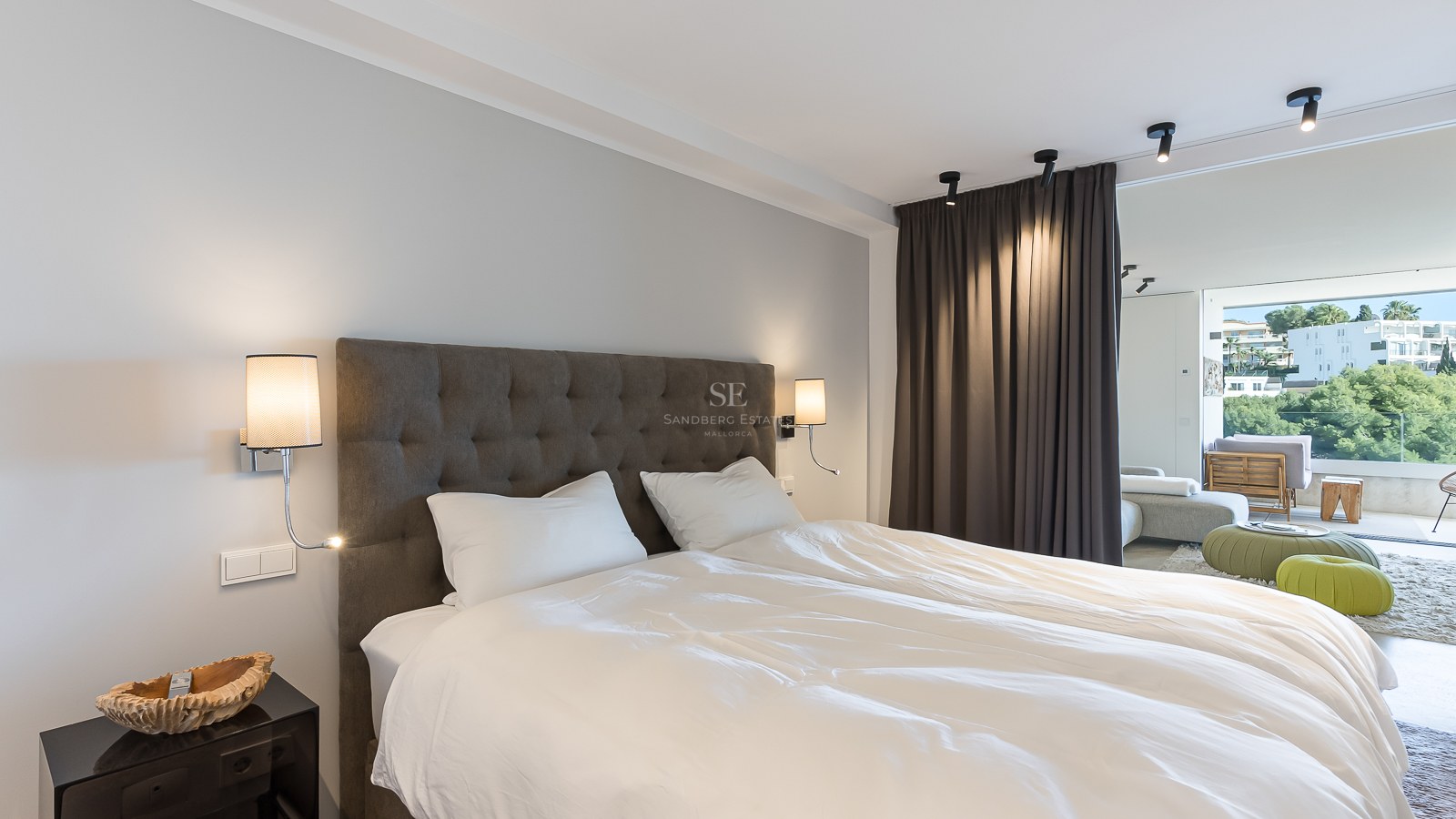 Chambre de maître moderne avec tête de lit capitonnée grise, draps blancs et baies vitrées ouvrant sur un balcon.