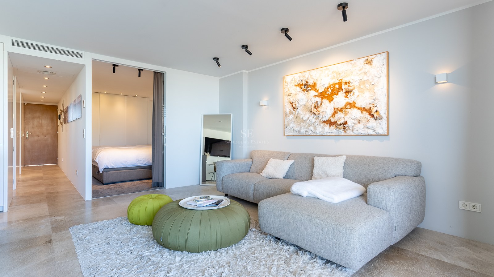 Salon lumineux avec canapé gris, poufs verts et tapis shaggy, offrant une vue sur la chambre attenante.