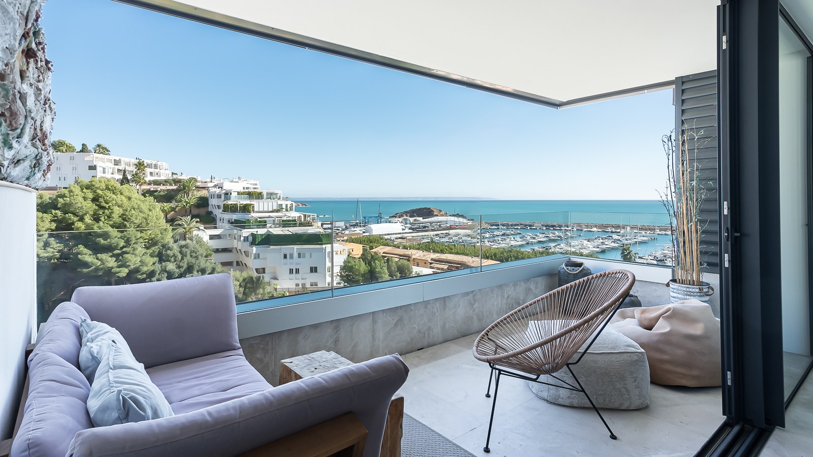 Terrasse couverte avec canapé lavande, fauteuil en osier et garde-corps en verre surplombant la marina et la mer.
