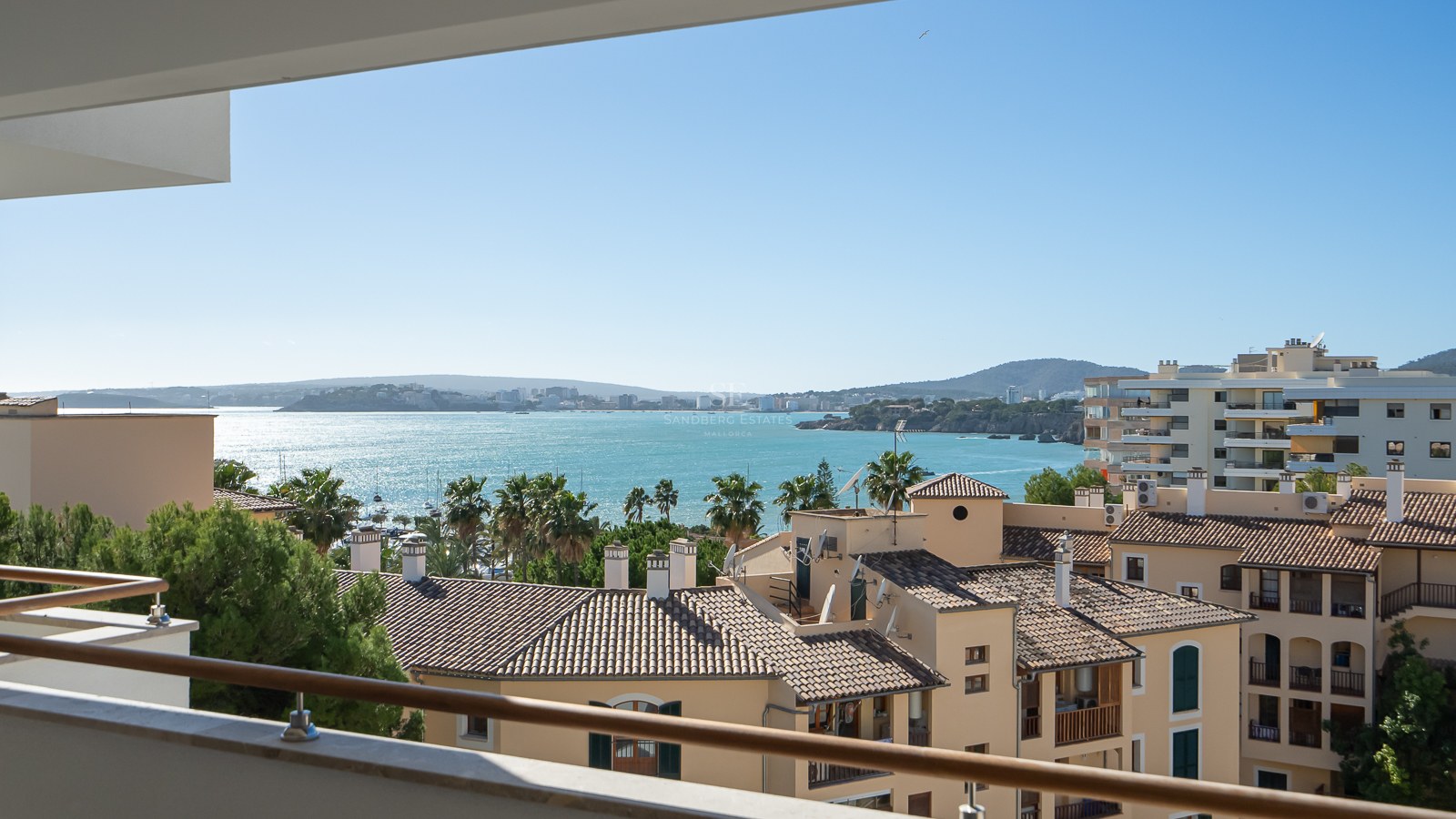 Vue d'un balcon surplombant des toits méditerranéens, des palmiers et une vaste baie turquoise sous un ciel bleu clair.