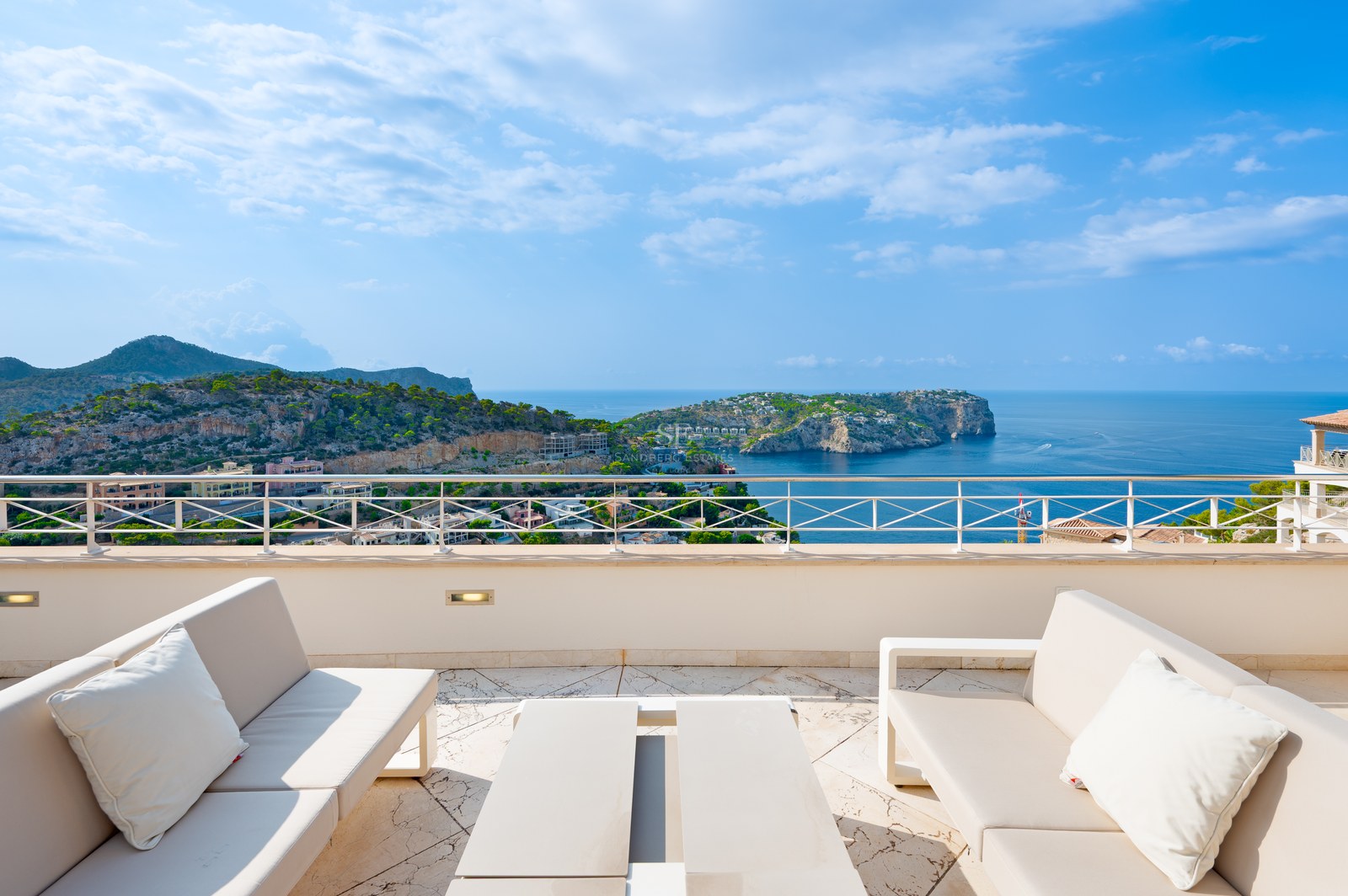 Elevated terrace with white outdoor sofas and a metal railing overlooking a blue bay and rocky Mediterranean coastline.