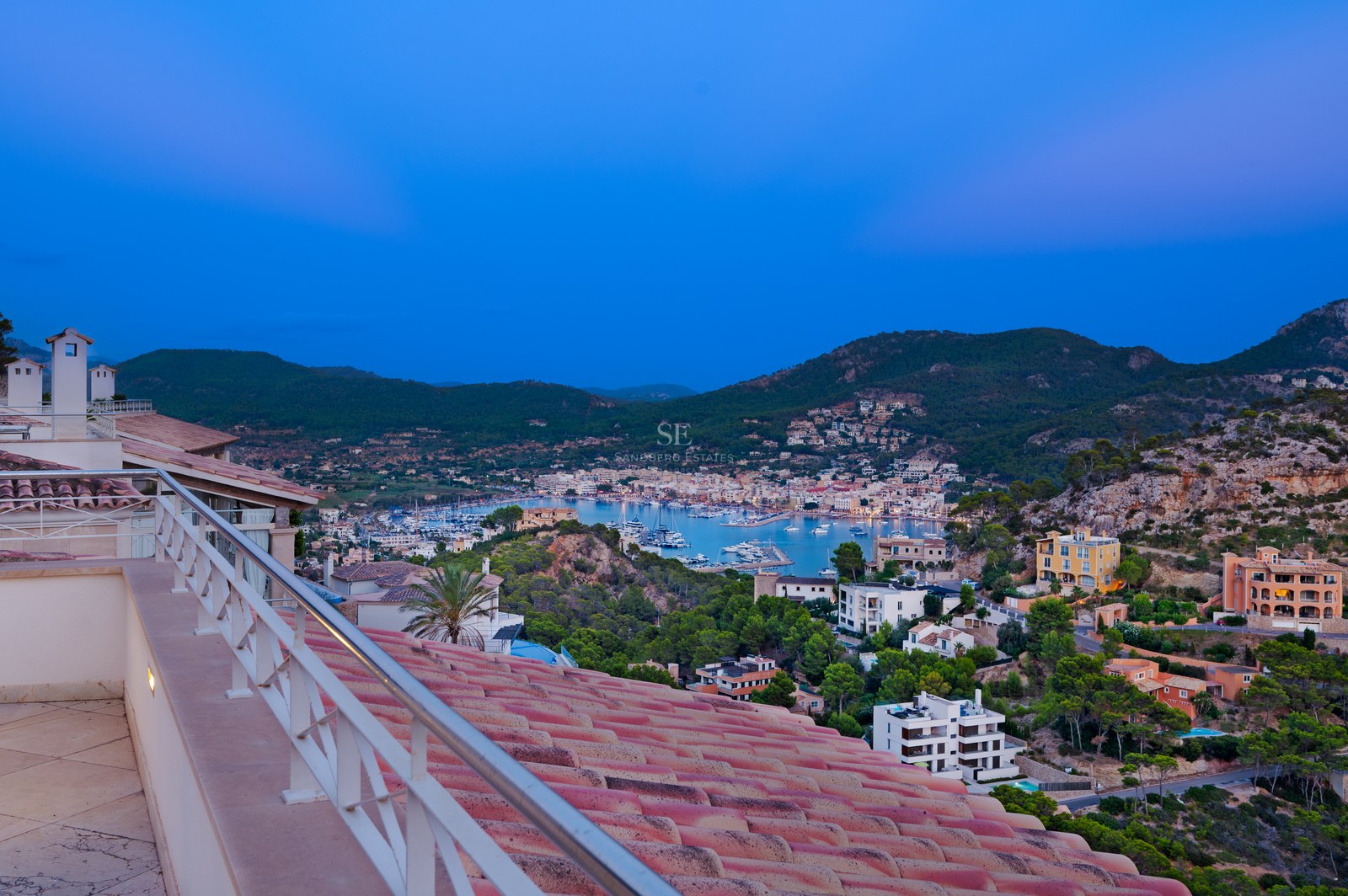 Elevated evening view of a Mediterranean harbor with boats, terracotta roofs in foreground, and mountains beyond.