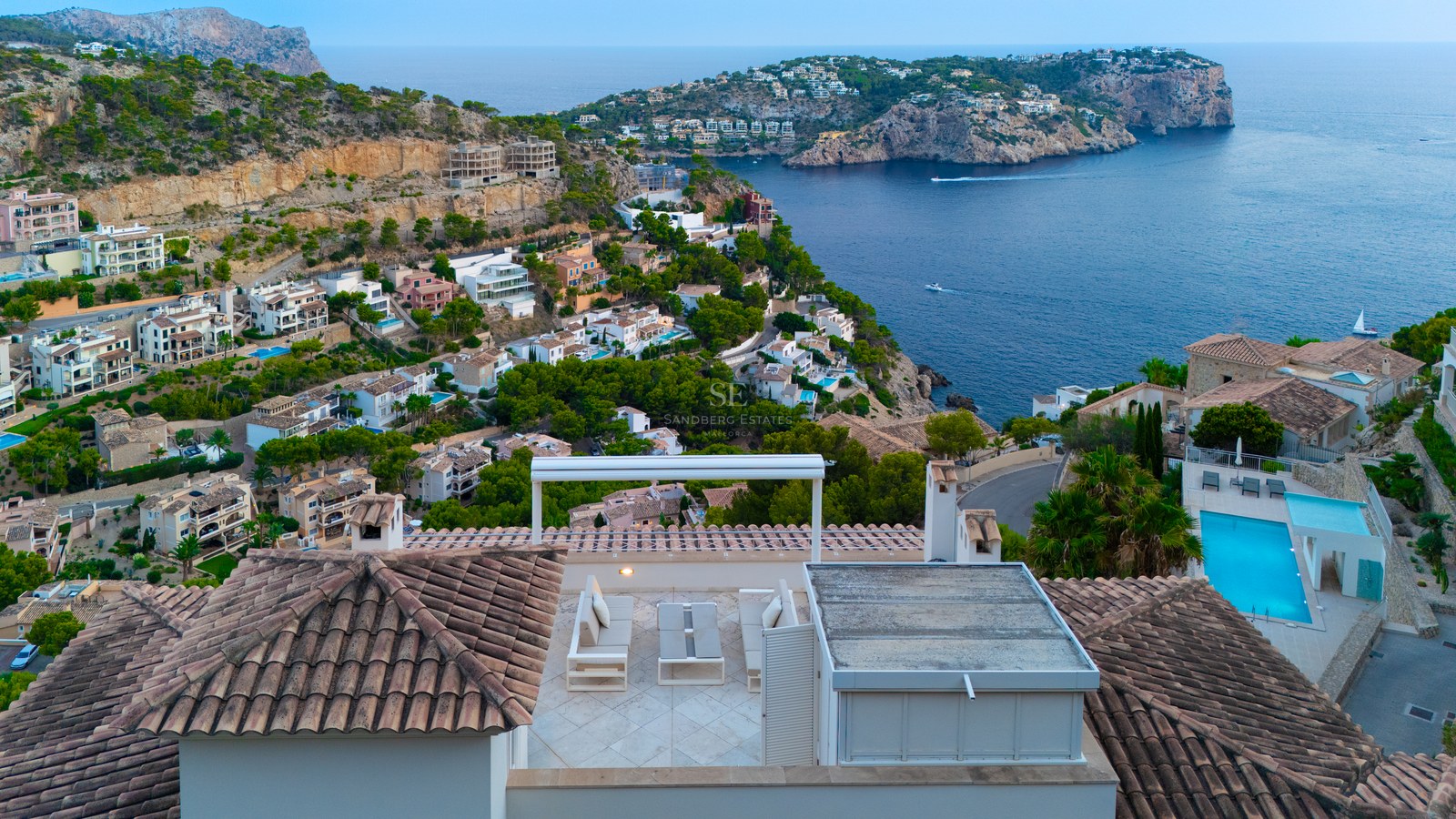 Elevated view of a white modern rooftop lounge with sofas overlooking a Mediterranean bay and coastal hills.
