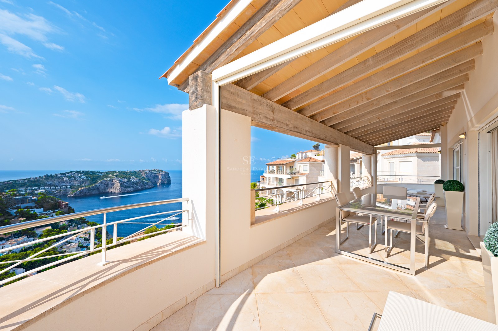 A sunny covered terrace with wooden beams and a dining area overlooking a blue sea and rocky coastline.