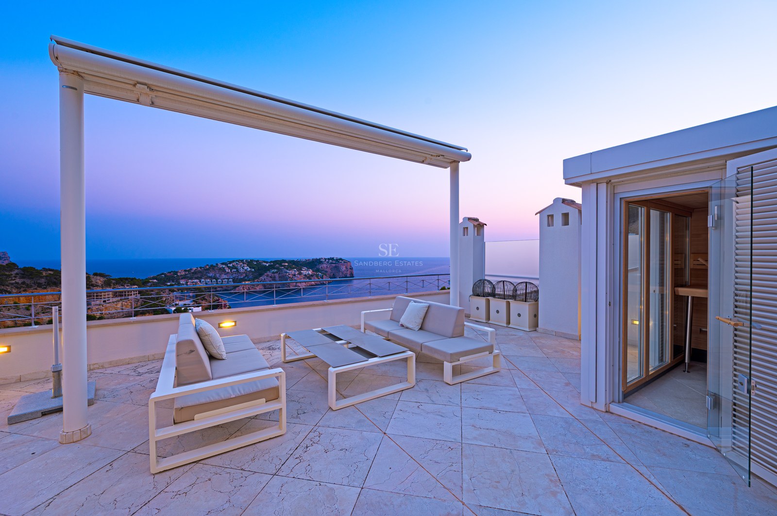 Modern rooftop terrace with white outdoor sofas overlooking a coastline at sunset.