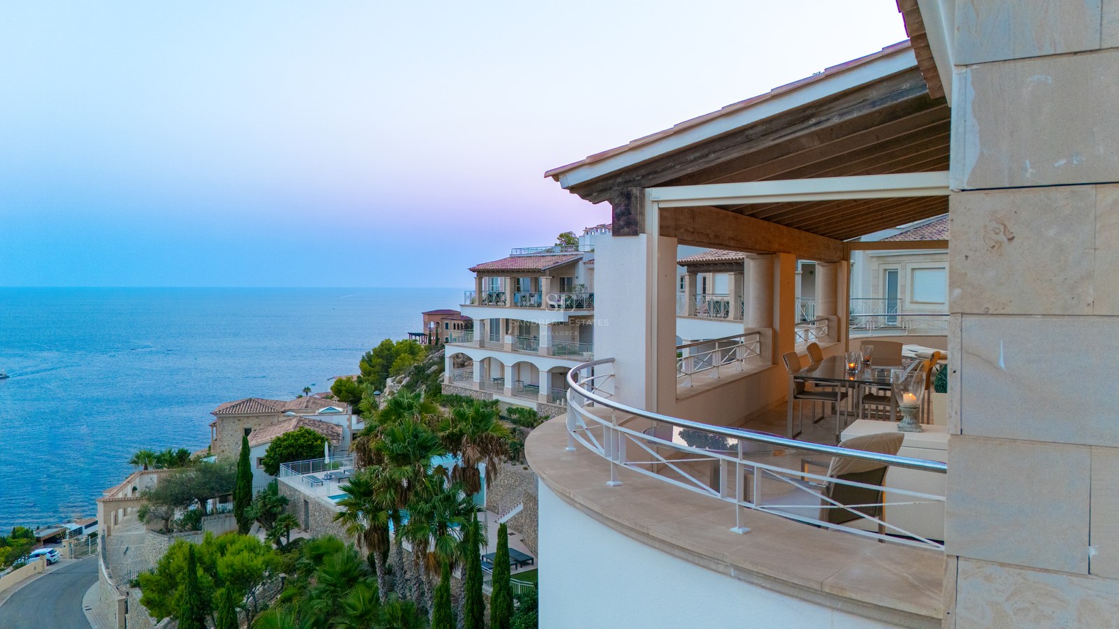 Curved stone balcony with stainless steel railings overlooking Mediterranean villas and the sea at sunset.