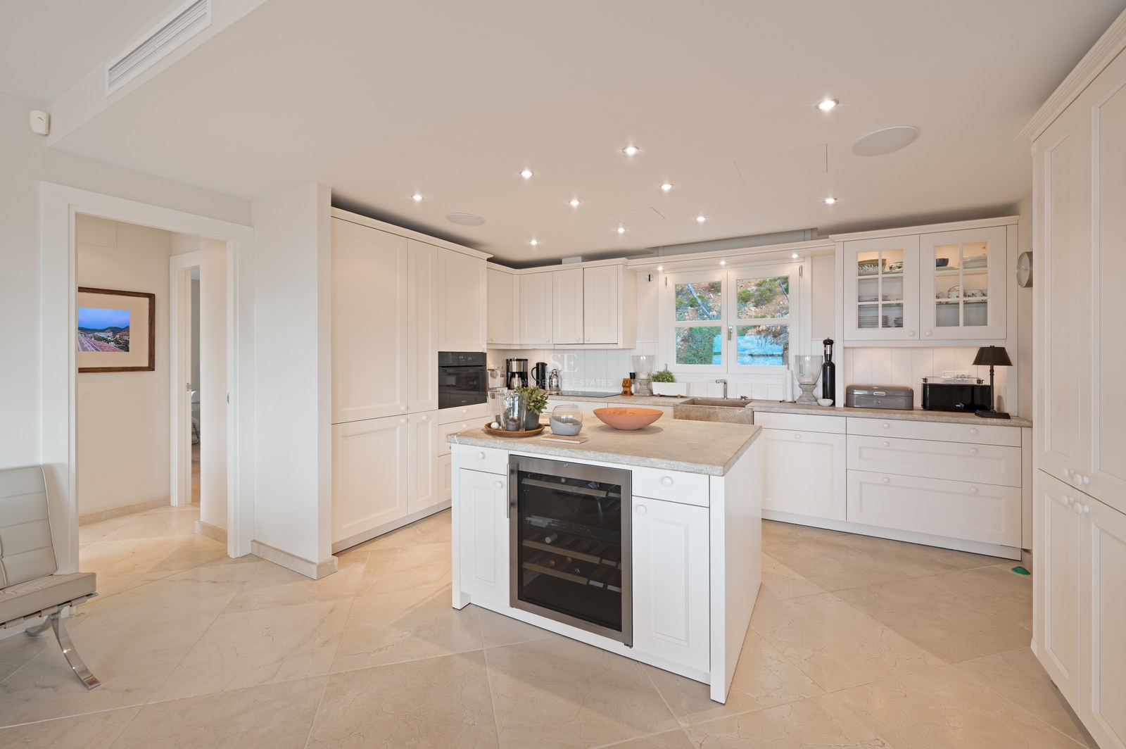 Bright white kitchen featuring a central island with integrated wine fridge, stone flooring, and recessed LED lighting.