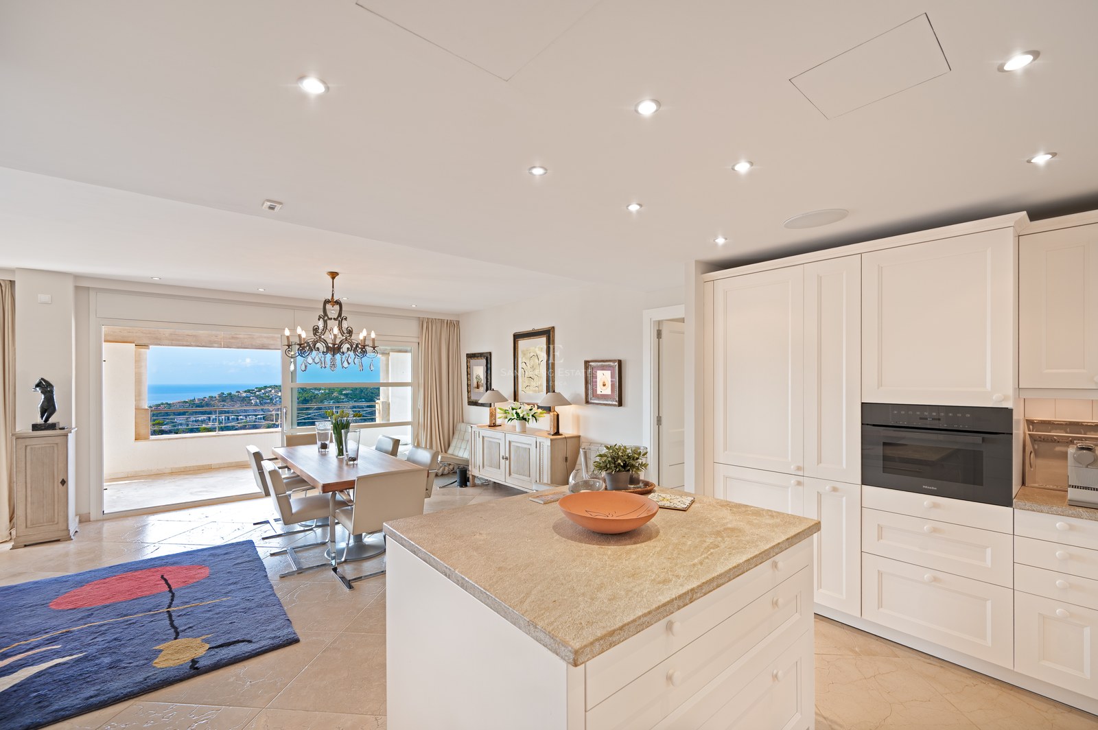 Modern white kitchen island and dining table in a bright room overlooking the sea through glass sliding doors.