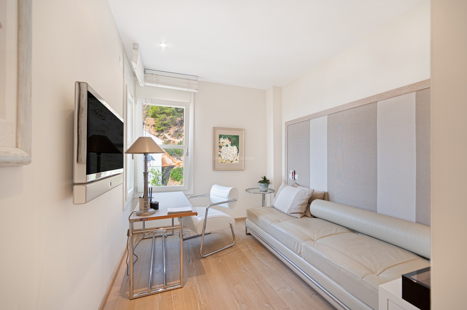 Bright home office featuring light wood floors, white leather sofa, glass desk, and a window showing greenery.
