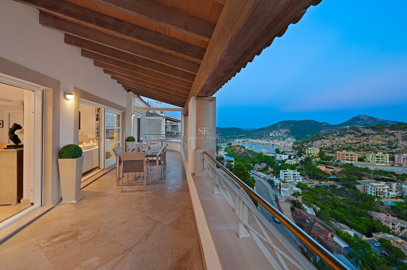 A spacious covered terrace with wooden ceiling beams, stone flooring, and a dining set overlooking a coastal town at dusk.