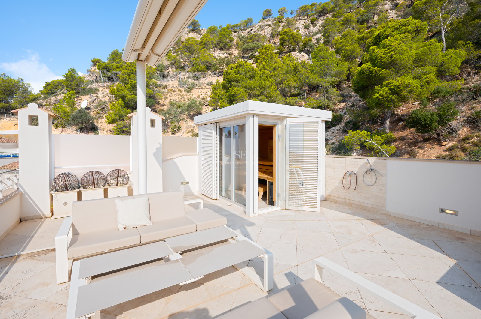 A sunny white stone terrace featuring an outdoor sauna cabin, lounge seating, and a backdrop of pine trees and rocky hills.