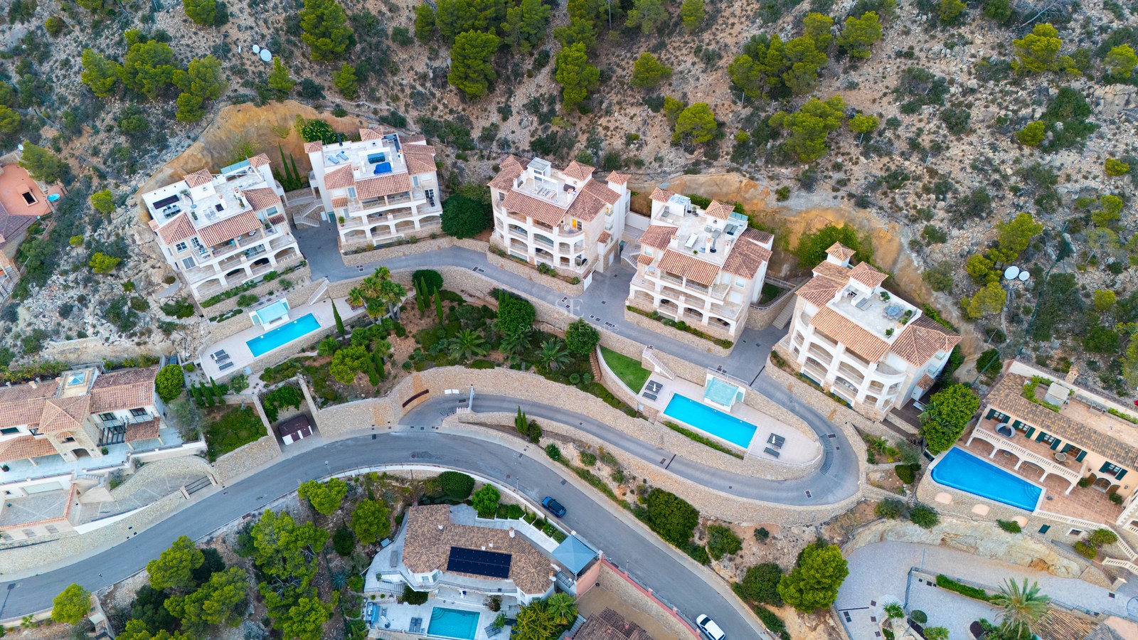 Overhead drone shot of luxury apartments with terracotta roofs and turquoise pools nestled in a green hillside.