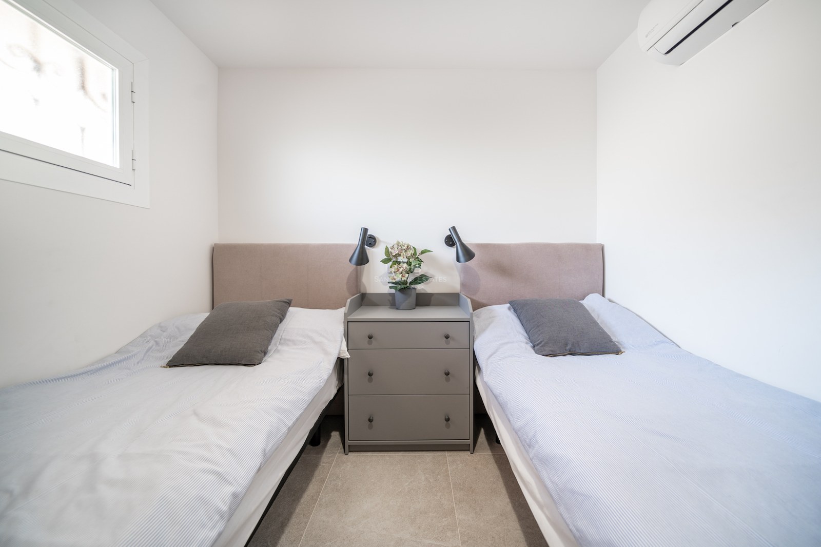 Chambre lumineuse avec deux lits simples, têtes de lit beiges, commode grise centrale et liseuses murales noires.