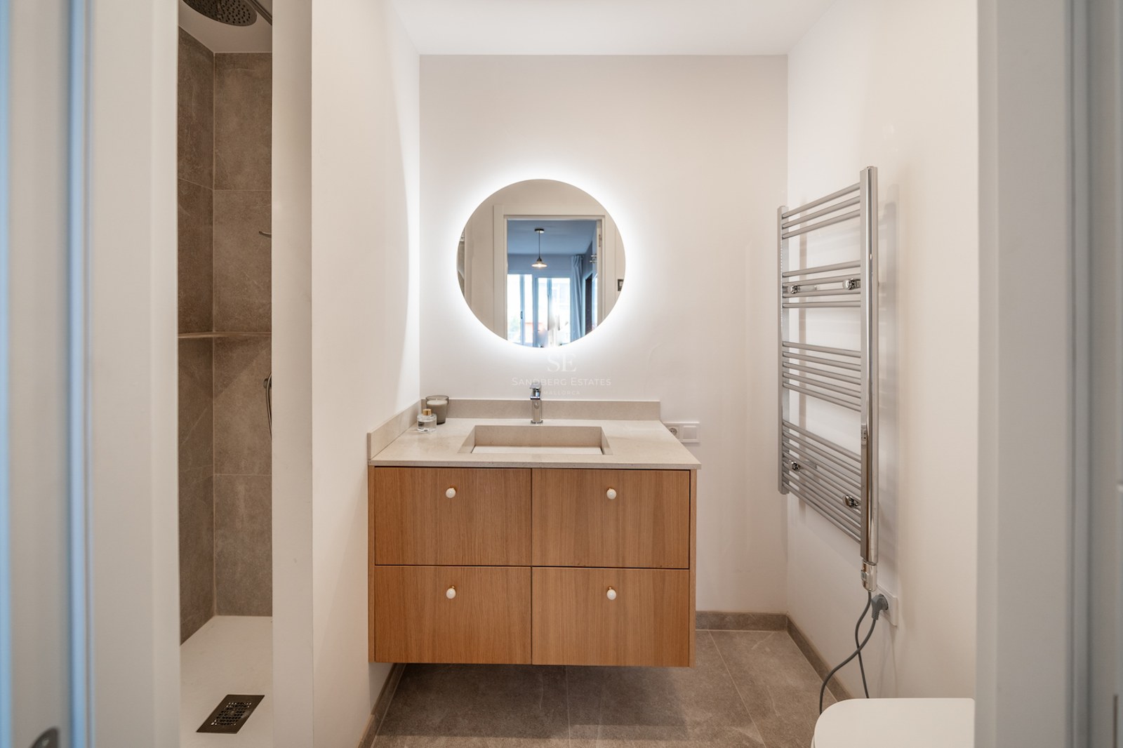 Salle de bain minimaliste avec miroir circulaire rétroéclairé, meuble en chêne, douche à l'italienne et sèche-serviettes.