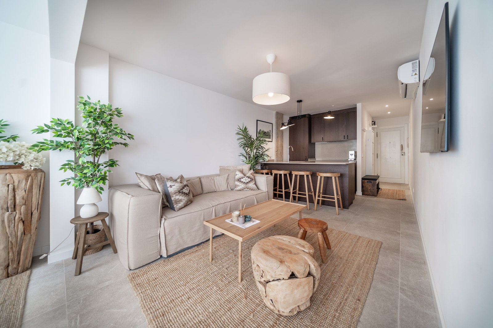 Salon lumineux à aire ouverte avec canapé beige, table basse en bois, tapis en jute et comptoir de cuisine.