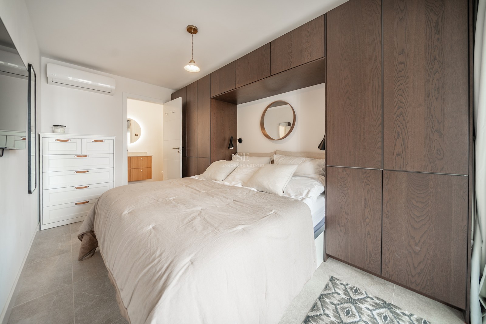 Chambre avec lit double, tête de lit encastrée dans des armoires en bois sombre et miroir rond décoratif.
