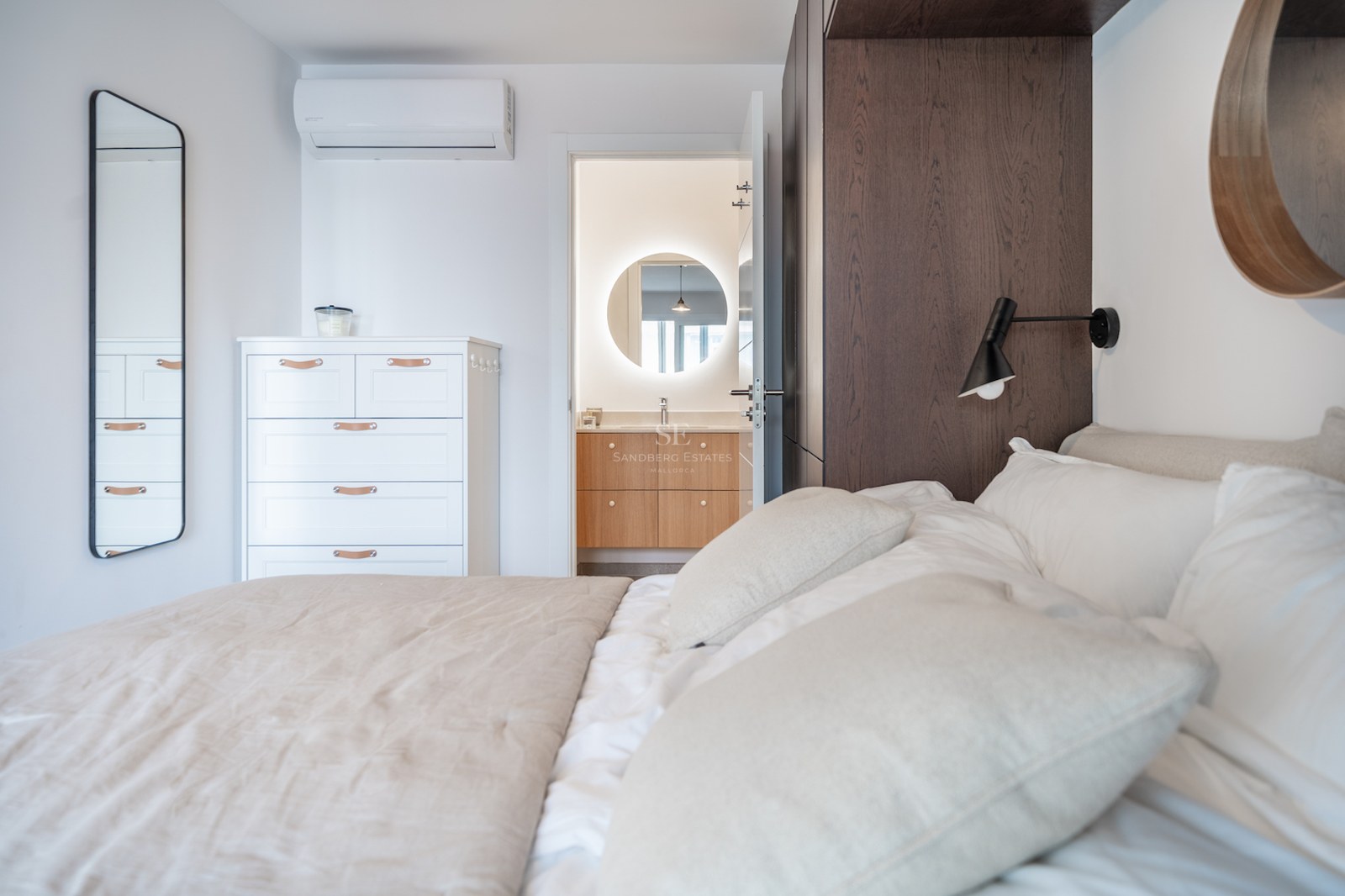 Chambre à coucher avec lit blanc ouvrant sur une salle de bain avec miroir rond rétroéclairé et meuble en bois.