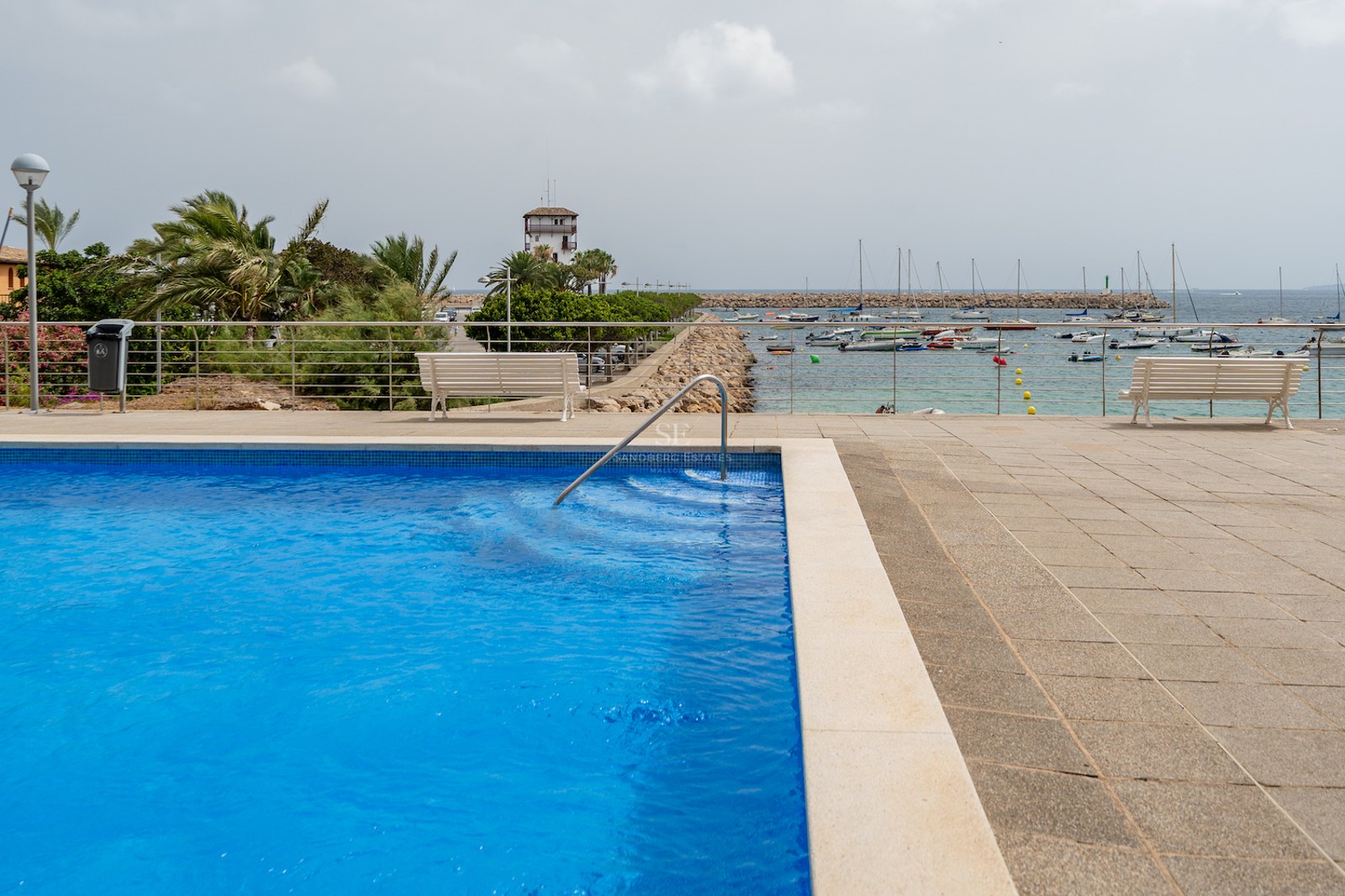Piscine bleue avec rampe en inox donnant sur un port de plaisance avec bateaux et promenade en pierre.